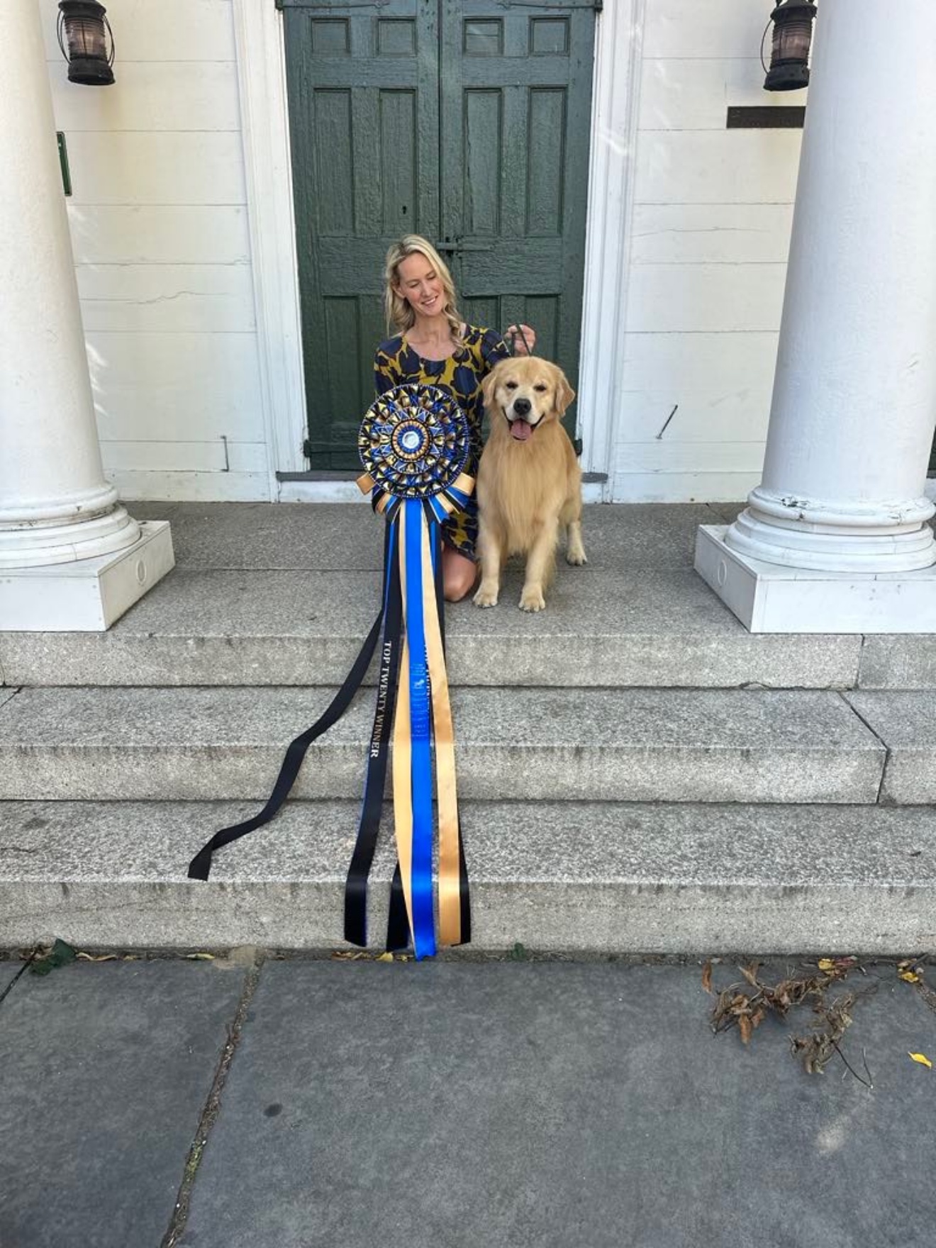 Amanda kneeling on stone steps beside a Golden Retriever with a large Top Twenty Winner rosette