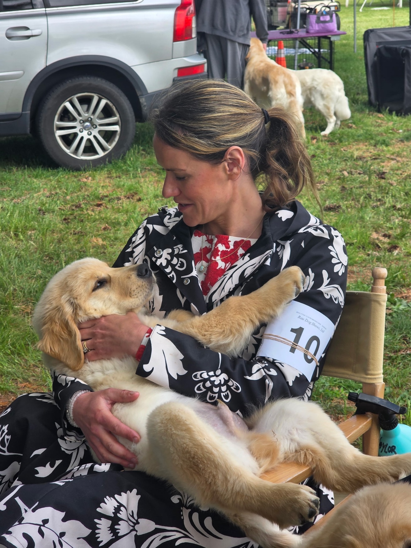 Amanda seated ringside at an outdoor dog show, exhibitor armband on, holding a Golden Retriever puppy against her face